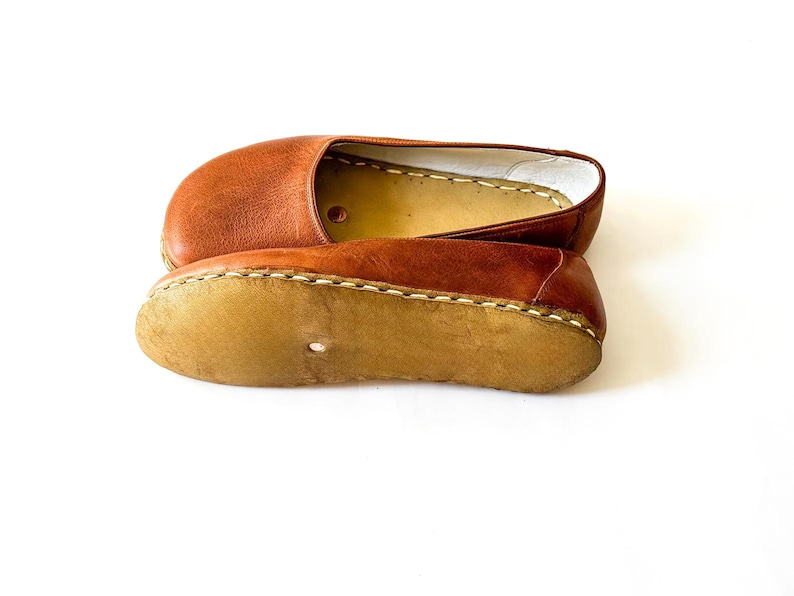 May include: A pair of tan leather slip-on shoes featuring a rounded toe, a smooth upper surface, and a light-colored, textured sole attached with visible thick stitching. The interior lining is a light cream color with a small circular cutout.