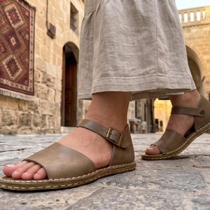 Op de afbeelding: Een paar taupekleurige leren sandalen met een brede voorband, een verstelbare enkelband met een messingkleurige metalen gesp en zichtbare lichtgekleurde stiksels langs de rand van de platte zool, gedragen met een beige linnen rok.