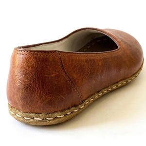 May include: A slip-on shoe crafted from textured brown leather with a rounded toe and a low-profile silhouette. The shoe features a thick, light-colored sole attached with visible, heavy-duty white stitching that runs along the entire perimeter.