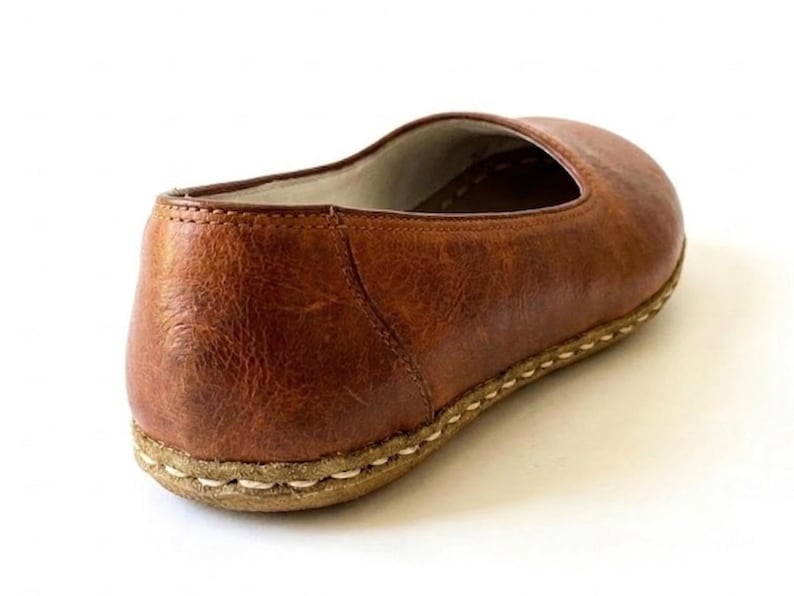 May include: A slip-on shoe crafted from textured brown leather with a rounded toe and a low-profile silhouette. The shoe features a thick, light-colored sole attached with visible, heavy-duty white stitching that runs along the entire perimeter.