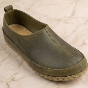 May include: An olive green slip-on shoe featuring a smooth leather-like upper with a center seam, a rounded toe, a pull tab at the heel, and a light-colored textured sole with visible stitching around the perimeter of the base of the shoe.