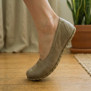May include: A taupe-colored leather flat shoe with a rounded toe and a textured, wrinkled surface. The shoe features a visible whipstitch detail along the sole edge, connecting the upper material to the base, providing a distinct decorative border.