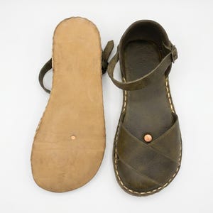 Peut inclure: Une paire de sandales en cuir vert olive avec des brides crois&eacute;es, une bride de cheville r&eacute;glable avec boucle en m&eacute;tal et une semelle plate beige. Les sandales pr&eacute;sentent des coutures apparentes de couleur claire sur les bords et un petit rivet cuivr&eacute; sur la bride avant.