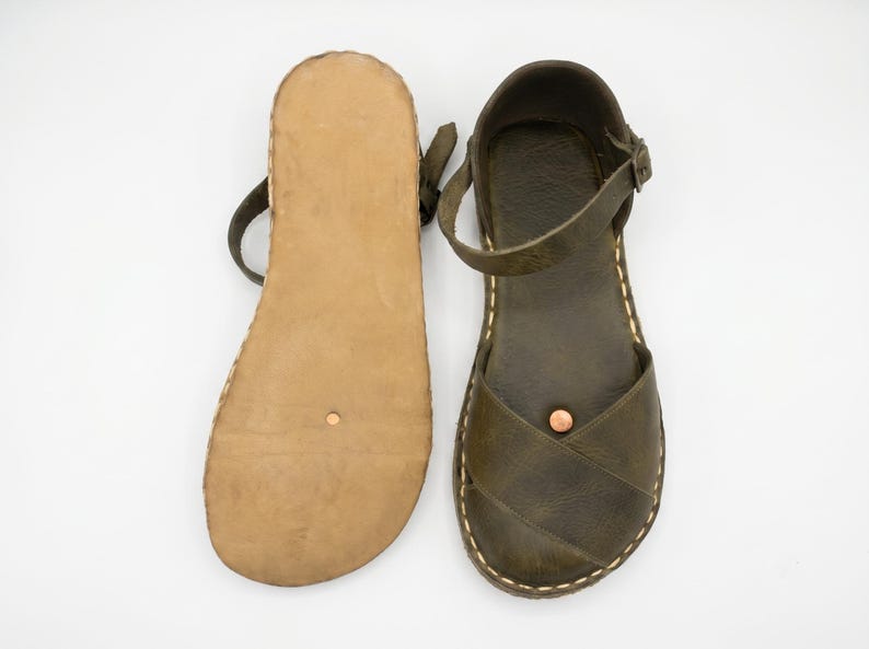 Peut inclure: Une paire de sandales en cuir vert olive avec des brides crois&eacute;es, une bride de cheville r&eacute;glable avec boucle en m&eacute;tal et une semelle plate beige. Les sandales pr&eacute;sentent des coutures apparentes de couleur claire sur les bords et un petit rivet cuivr&eacute; sur la bride avant.