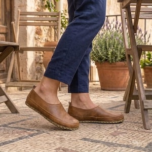 Puede incluir: Zapatos sin cordones de cuero marr&oacute;n con punta redondeada. Los zapatos tienen una suela cosida y se usan con pantalones azul marino. La imagen est&aacute; tomada al aire libre, con sillas de madera y plantas en macetas en el fondo.