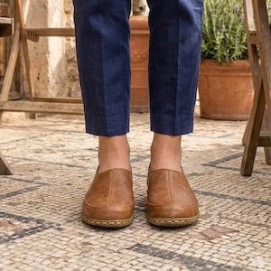 Puede incluir: Un par de zapatos sin cordones de cuero marr&oacute;n con punta redondeada. Los zapatos se usan con pantalones azul marino. Los zapatos tienen una suela cosida y est&aacute;n sobre un suelo de baldosas de mosaico. Sillas de madera y plantas en macetas en el fondo.