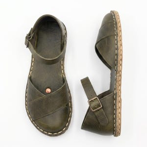 Peut inclure: Une paire de sandales en cuir vert olive avec un design &agrave; brides crois&eacute;es sur le dessus, une bride de cheville r&eacute;glable avec boucle en m&eacute;tal, un petit rivet couleur cuivre sur la semelle et des coutures claires visibles le long de la semelle.