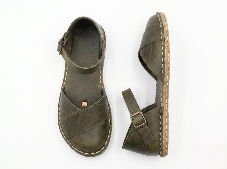 Peut inclure: Une paire de sandales en cuir vert olive avec un design &agrave; brides crois&eacute;es sur le dessus, une bride de cheville r&eacute;glable avec boucle en m&eacute;tal, un petit rivet couleur cuivre sur la semelle et des coutures claires visibles le long de la semelle.
