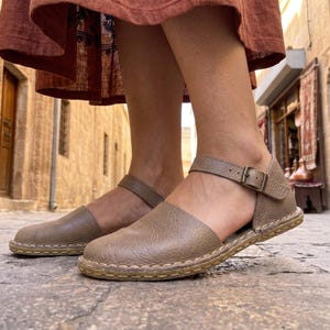 May include: Tan leather sandals with a buckle closure and ankle strap. The sandals have a stitched sole and are worn outdoors on a cobblestone street. The sandals are a neutral color and appear to be comfortable.