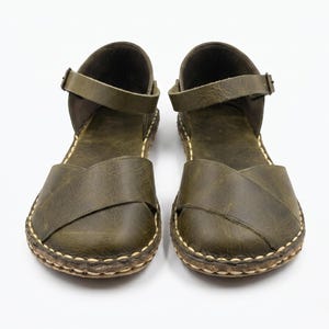 Peut inclure: Une paire de sandales en cuir vert olive dot&eacute;es d'une large bride crois&eacute;e sur l'avant-pied, d'une bride de cheville r&eacute;glable avec une petite boucle en m&eacute;tal et d'une semelle &eacute;paisse avec des surpiq&ucirc;res contrastantes claires sur le bord.