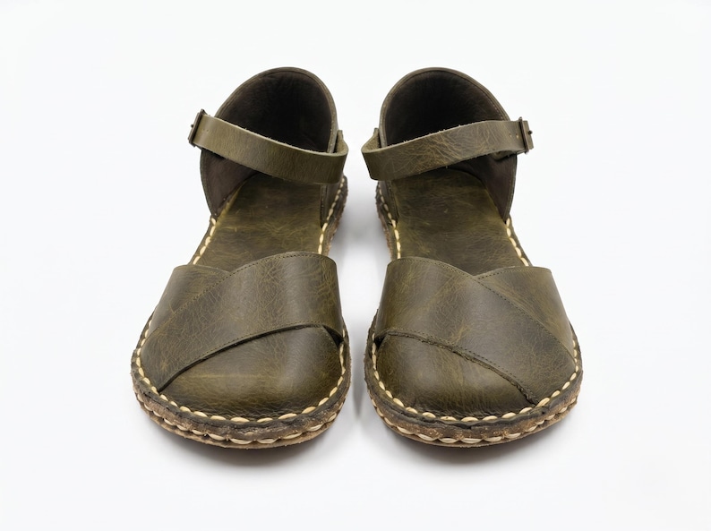 Peut inclure: Une paire de sandales en cuir vert olive dot&eacute;es d'une large bride crois&eacute;e sur l'avant-pied, d'une bride de cheville r&eacute;glable avec une petite boucle en m&eacute;tal et d'une semelle &eacute;paisse avec des surpiq&ucirc;res contrastantes claires sur le bord.