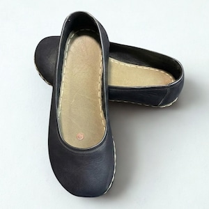 May include: A pair of dark navy blue slip-on flats featuring a rounded toe, a smooth exterior finish, and a light beige interior insole. The shoes are accented with visible white stitching along the perimeter of the sole and a small circular logo.