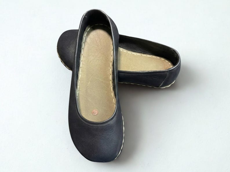 May include: A pair of dark navy blue slip-on flats featuring a rounded toe, a smooth exterior finish, and a light beige interior insole. The shoes are accented with visible white stitching along the perimeter of the sole and a small circular logo.