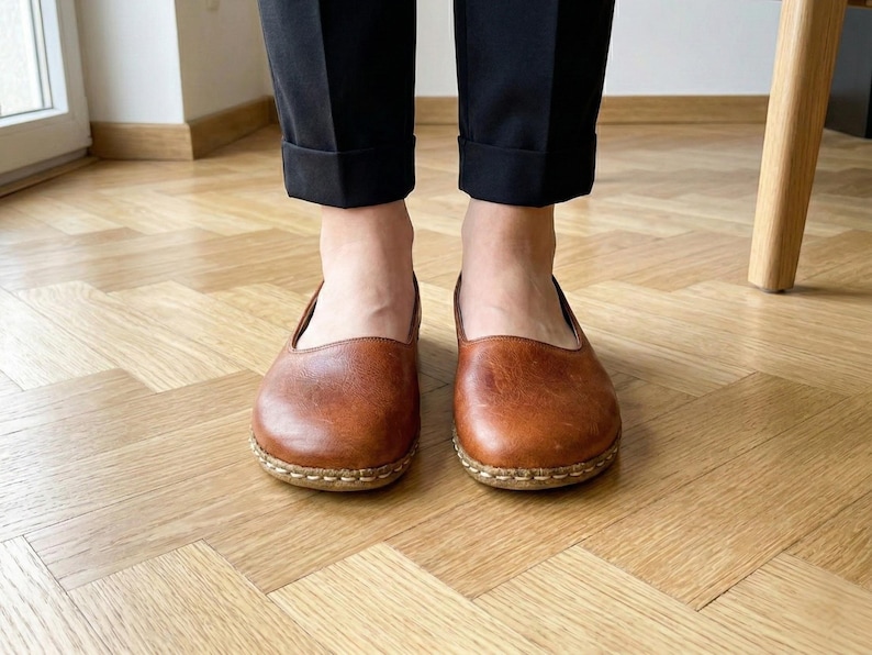 May include: A pair of brown leather slip-on shoes featuring a rounded toe, a low-cut topline, and visible light-colored stitching along the sole edge. The shoes are worn with black cuffed trousers, showcasing a simple and functional design.