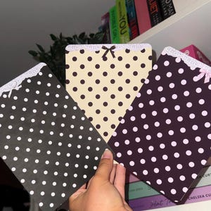 Può includere: Tre blocchi per appunti a pois neri, crema e viola scuro. Ogni blocco per appunti ha un bordo in pizzo bianco e un piccolo fiocco. I blocchi per appunti sono tenuti davanti a una libreria con titoli di libri visibili.