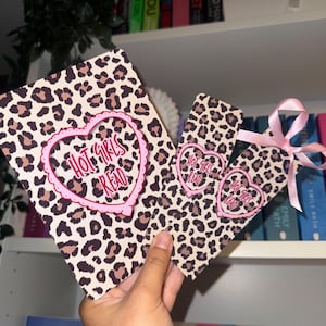 May include: A rectangular card and a matching bookmark, both featuring a tan and brown leopard print pattern. Each item displays a pink heart-shaped frame containing the text "HOT GIRLS READ". The bookmark is finished with a light pink ribbon.