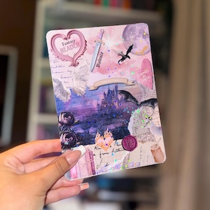 May include: A decorative card with a fantasy theme. It features a heart-shaped frame with the words "Fantasy Reader", a sword labelled "Violence", a bat, and the words "Enemies to Lovers". The colour scheme includes pink, purple, and white.