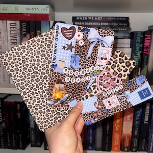 Könnte beinhalten: Eine Hand hält mehrere dekorative Lesezeichen. Die Lesezeichen zeigen ein Leopardenmuster, Schleifen und den Text "One more chapter" und "Probably Reading Books". Weitere Designs sind Stiefel, Kirschen und Sterne. Im Hintergrund ist ein Bücherregal mit Büchern zu sehen.