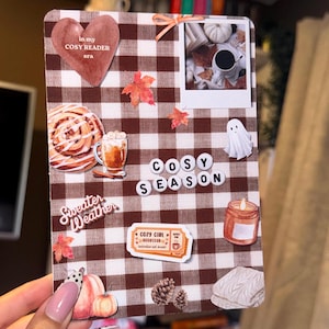 May include: A rectangular card with a brown and white gingham pattern featuring stickers of a heart with text in my COSY READER era, a polaroid, a cinnamon roll, a ghost, a candle, pumpkins, pinecones, and text reading COSY SEASON.