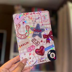 May include: A decorative notebook with a pink and white checkered pattern, adorned with cowboy-themed stickers. Features a pink cowboy hat, horseshoe, star with "Wild West" text, dice, playing cards, and the text "Howdy".