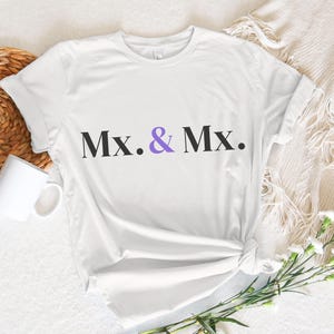 Op de afbeelding: Wit T-shirt met de tekst "Mx. & Mx." in zwart en paars. Het shirt is in de taille geknoopt en ligt op een wit oppervlak. Een witte mok en een gevlochten mand zijn links zichtbaar. Rechts is een boeket witte bloemen.