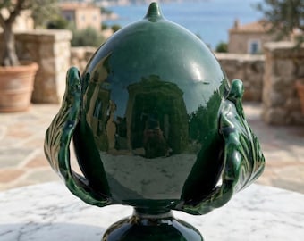 May include: A glossy dark green ceramic decorative object shaped like a pinecone with a pointed top, resting on a circular pedestal base. The piece features sculpted leaf-like details on both sides and has a smooth, reflective glazed finish.