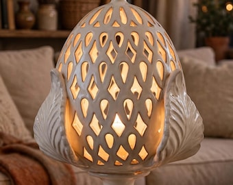 May include: A cream-colored ceramic lamp shaped like a pinecone with an intricate lattice cutout pattern. The lamp features a pedestal base and decorative leaf-like accents at the bottom, emitting a warm glow from an internal light source.
