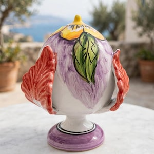 Könnte beinhalten: Ein dekoratives Objekt aus Keramik in Form eines Tannenzapfens mit weißem Untergrund, violetten Pinselstrichen, einer gelben Blume an der Spitze, einem grünen Blatt und zwei auffälligen roten, gewellten, flügelartigen Seitenelementen.