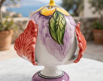May include: A ceramic pinecone-shaped decorative object featuring a white base with hand-painted purple brushstrokes, a yellow flower at the top, a green leaf, and two prominent red, ruffled, wing-like side elements on a purple pedestal base.