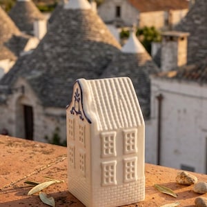Può includere: Una statuetta di casa in miniatura in ceramica bianca con tetto a capanna, pareti con trama a mattoni e dettagli delle finestre in rilievo. La facciata anteriore presenta un emblema decorativo dipinto in blu sulla sommità.
