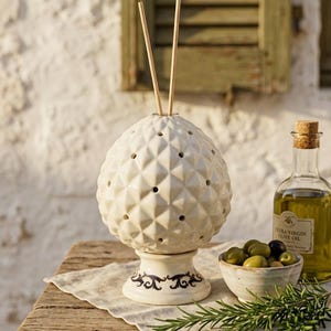 Può includere: Un diffusore in ceramica bianca con una superficie a motivo geometrico a rombi e piccoli fori circolari, poggiante su una base con accenti decorativi blu scuro. Due sottili bastoncini di legno sporgono dalla parte superiore del contenitore.