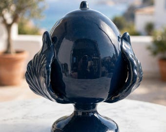 May include: A glossy, dark navy blue ceramic decorative object shaped like a pinecone or egg, featuring a central rounded body with a small pointed top, supported by a pedestal base and flanked by two symmetrical, leaf-like side ornaments.
