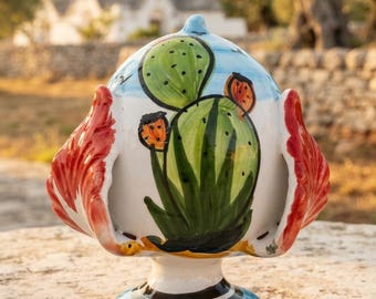 May include: A ceramic pumo ornament featuring a hand-painted green cactus with orange flowers on a white background. The object has a rounded top, a flared base with blue and black stripes, and two large, red, leaf-like side protrusions.
