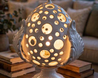 May include: A white ceramic table lamp with a rounded, dome-shaped body featuring numerous circular cutouts of varying sizes. The lamp is supported by a pedestal base and is flanked by two sculpted leaf-like elements on either side.