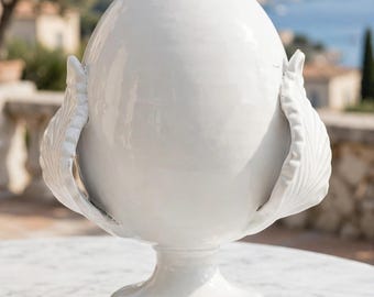 May include: A glossy white ceramic decorative object shaped like an egg, featuring a smooth central dome supported by a pedestal base and flanked by two symmetrical, leaf-like structures with ribbed textures on either side.