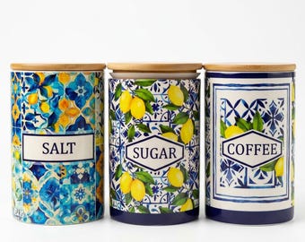 May include: Three cylindrical ceramic canisters with light wood lids, each featuring a blue and white tile pattern with yellow lemon motifs. The canisters are labeled with black text reading SALT, SUGAR, and COFFEE in rectangular white frames.