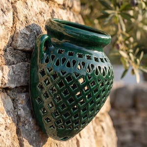 May include: A dark green glazed ceramic wall planter with a wide, rounded rim and a flat back for mounting. The body features a decorative lattice pattern with repeating geometric cutouts and a single handle on the side, creating an openwork design.
