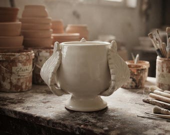 May include: A white ceramic vessel with a smooth, glossy finish, featuring two prominent, wing-shaped handles on the sides. The vessel has a wide, rounded body that tapers down to a circular pedestal base, centered on a worn, textured workbench.