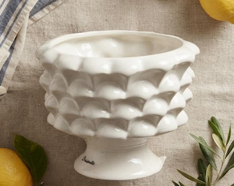 May include: A white ceramic bowl with a textured, scalloped exterior pattern and a pedestal base. The word "Oriente" is printed in small black script on the side of the base. The bowl has a wide, open top and a glossy, smooth finish.