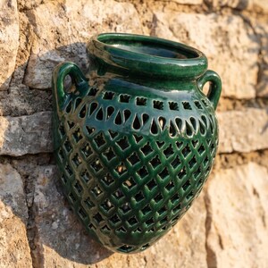 May include: A dark green glazed ceramic wall planter featuring a rounded, amphora-like shape with two side handles. The body of the vessel is decorated with a repeating lattice pattern of cut-out diamond shapes and a row of arched openings at the top.