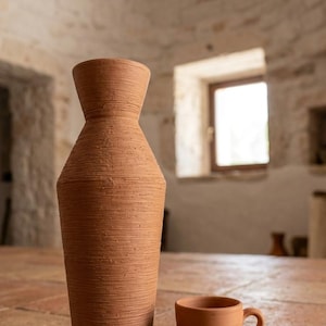 Peut inclure: Un grand vase en terre cuite élancé avec une surface texturée et nervurée et un col étroit, placé à côté d'une petite tasse en terre cuite lisse avec une anse. Les deux objets présentent une finition en argile brun rougeâtre naturelle.