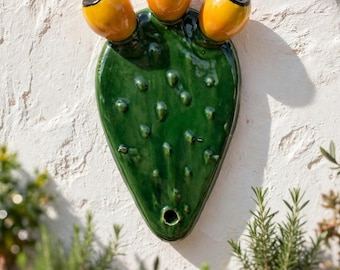 May include: A glossy green ceramic wall sculpture shaped like a prickly pear cactus pad. The piece features three rounded yellow fruits with black centers at the top and a small circular opening at the bottom, all finished with a smooth glaze.