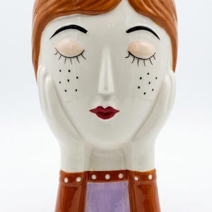 Può includere: Un vaso in ceramica a forma di testa umana con occhi chiusi, ciglia lunghe e lentiggini. La figura ha capelli castano-rossicci, labbra rosse e mani appoggiate sulle guance. La base presenta una maglia viola con una giacca marrone e pois bianchi.