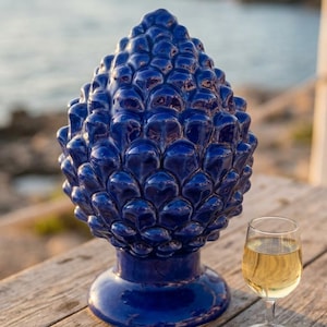 Può includere: Una scultura in ceramica a forma di pigna di colore blu profondo e lucido, con una base arrotondata e un motivo a scaglie sovrapposte. Accanto alla scultura si trova un piccolo bicchiere trasparente contenente un liquido giallo pallido.