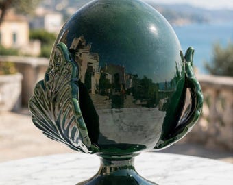 May include: A dark green ceramic vessel with a glossy finish, featuring a rounded central body and a pedestal base. The object is adorned with sculpted leaf-like elements on both sides, reflecting the surrounding outdoor environment.