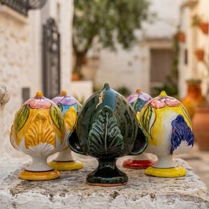 Può includere: Una collezione di cinque ornamenti in ceramica a forma di pigna, ciascuno dotato di una base a piedistallo. Il pezzo centrale è verde scuro con dettagli a foglia, mentre i pezzi circostanti sono dipinti con vivaci motivi floreali gialli, bianchi, rosa e blu.