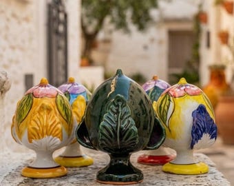 May include: A collection of five ceramic pinecone-shaped ornaments, each featuring a pedestal base. The central piece is dark green with leaf detailing, while the surrounding pieces are painted in vibrant yellow, white, pink, and blue floral patterns.