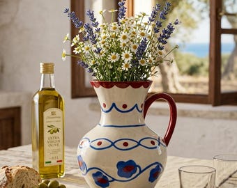 May include: A cream-colored ceramic pitcher with a red rim and handle, decorated with blue and red hand-painted floral and wavy line patterns. The pitcher is filled with a bouquet of purple lavender and white daisies, standing on a wooden table.