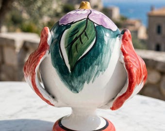 May include: A hand-painted ceramic urn featuring a white base with a central purple flower and green leaf motif. The vessel has two prominent red, wing-like side handles and sits on a circular red pedestal base with a black accent line.