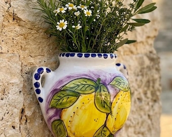 May include: A white ceramic wall planter with two handles, decorated with hand-painted yellow lemons, green leaves, and purple accents. The rim features a pattern of blue dots. The planter is filled with green herbs and small white daisy-like flowers.